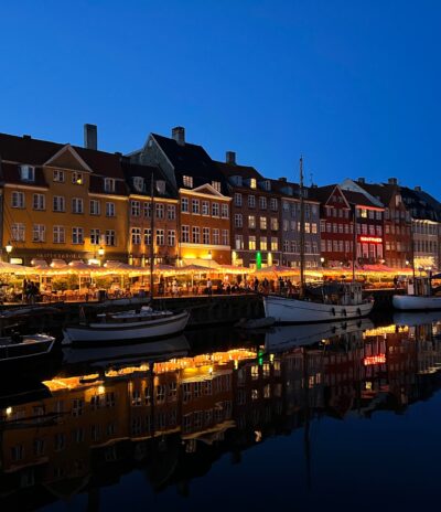 Kööpenhaminan kanava Hyhavn iltavalaistuksessa. Kuva joen toiselta puolelta, jossa etualalla veneitä, valaistuja ravintolaterasseja ja taustalla värikkäitä taloja.