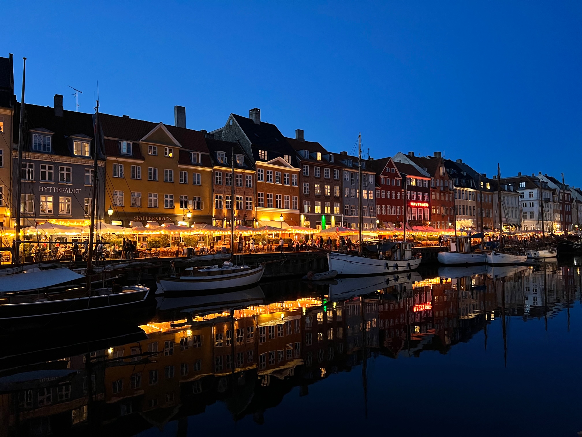 Kööpenhaminan kanava Hyhavn iltavalaistuksessa. Kuva joen toiselta puolelta, jossa etualalla veneitä, valaistuja ravintolaterasseja ja taustalla värikkäitä taloja.