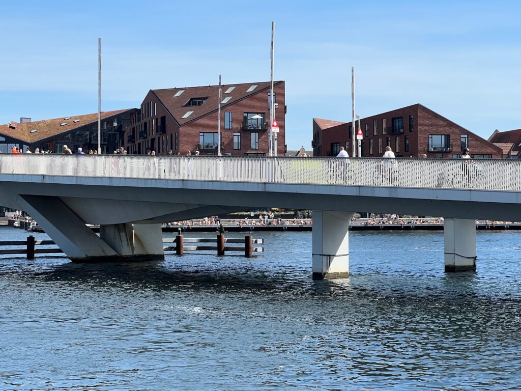 View to the other side of Nyhavn: a modern bridge over the canal with cyclists. In the back modern brick colored houses with geographical forms.